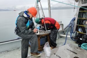 Two people work with sapling equipment aboard a vessel. An oil tanker can be seen in the background. 