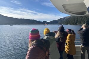 Several adults watch boats in the distance. The boats are a variety of smaller vessels, mostly fishing vessels, and one larger brightly colored vessel named the Ross Chouest. Lines of oil spill boom are attached to the smaller vessels in pairs, forming a "u" shape. 