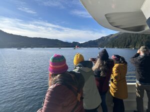 Several adults watch boats in the distance. The boats are a variety of smaller vessels, mostly fishing vessels, and one larger brightly colored vessel named the Ross Chouest. Lines of oil spill boom are attached to the smaller vessels in pairs, forming a "u" shape. 